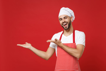 Cheerful young bearded male chef cook baker man in striped apron white t-shirt toque chefs hat posing isolated on red wall background. Cooking food concept. Mock up copy space. Pointing hands aside.