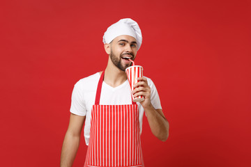 Handsome young bearded male chef cook or baker man in striped apron white t-shirt toque chefs hat isolated on red background. Cooking food concept. Mock up copy space. Holding cup of cola or soda.