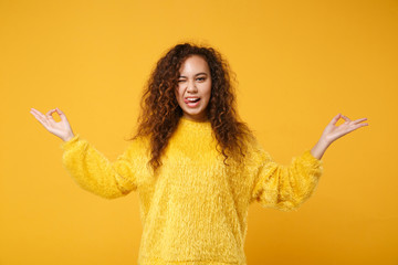 Funny african american girl in fur sweater posing isolated on yellow orange background. People lifestyle concept. Mock up copy space. Hold hands in yoga gesture relaxing meditating, showing tongue.