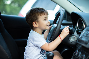 Little boy sitting behind the wheel of a car with a lovely smile on his face, outdoors, family travel concept