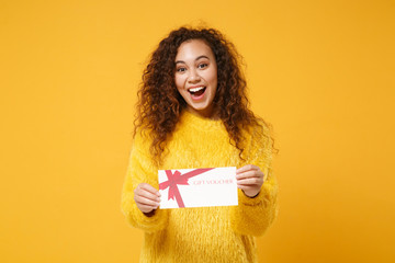 Excited young african american girl in fur sweater posing isolated on yellow orange background studio portrait. People lifestyle concept. Mock up copy space. Hold gift certificate, keeping mouth open.