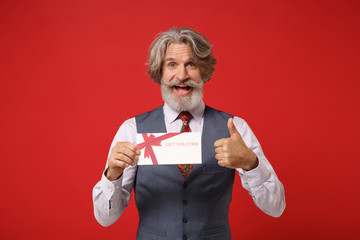 Cheerful elderly gray-haired mustache bearded man in classic shirt vest colorful tie isolated on red background. People lifestyle concept. Mock up copy space. Hold gift certificate showing thumb up.