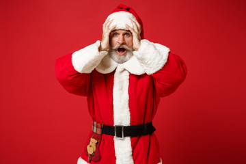 Fototapeta na wymiar Shocked elderly gray-haired mustache bearded Santa man in Christmas hat posing isolated on red background. New Year 2020 celebration holiday concept. Mock up copy space. Scream putting hands on head.