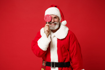 Elderly gray-haired mustache bearded Santa man in Christmas hat posing isolated on red wall background. New Year 2020 celebration holiday concept. Mock up copy space. Covering eye with round lollipop.