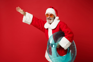 Fototapeta na wymiar Cheerful elderly gray-haired mustache bearded Santa man in Christmas hat sunglasses posing isolated on red background. New Year 2020 celebration concept. Mock up copy space. Holding inflatable ring.