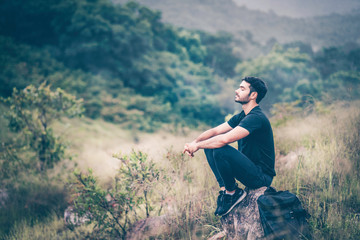 Young man sitting on natural stone in mountain, breathing fresh air after relaxing from hiking, feel free, freedom, refreshment and release. Walking in forest, camping activity lifestyle in summer.