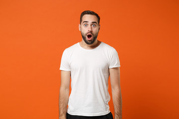 Shocked amazed young man in casual white t-shirt posing isolated on orange wall background, studio portrait. People sincere emotions lifestyle concept. Mock up copy space. Keeping mouth wide open.
