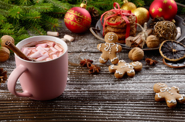 Christmas background of pink mug of hot chocolate with marshmallows, spruce branch and tray with gingerbread cookies on wooden table