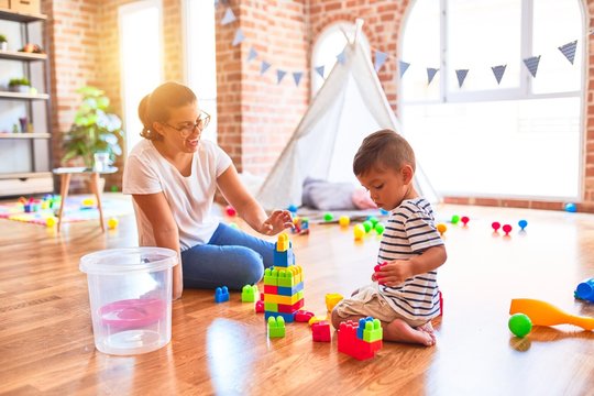 Beautiful teacher and toddler boy playing with construction blocks bulding tower at kindergarten