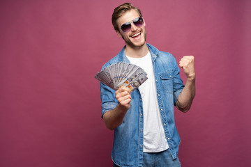 Joyful man in jeans shirt holding bunch of money banknotes while standing and celebrating isolated over pink background