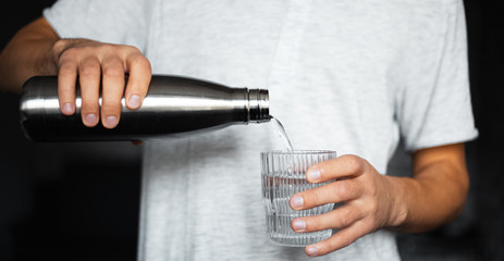 Male hands pours water into a glass from steel thermo eco bottle.