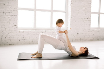 young sporty mother does physical yoga or pilates exercises, butt bridge, together with her baby against big windows background. Fitness, happy maternity and healthy lifestyle concept.