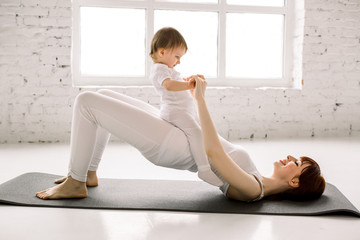 Young happy mother working out, doing butt bridge exercise, wearing white sportswear, little baby...