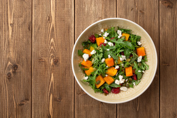 Autumn salad with baked pumpkin, arugula, seeds, dried cranberries and feta cheese in bowl on rustic wooden background. Top view. Copy space.
