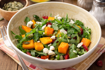 Autumn salad with baked pumpkin, arugula, seeds, dried cranberries and feta cheese in bowl on rustic wooden background. Selective focus.