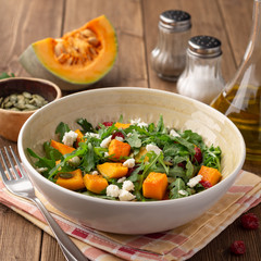 Autumn salad with baked pumpkin, arugula, seeds, dried cranberries and feta cheese in bowl on rustic wooden background. Selective focus.