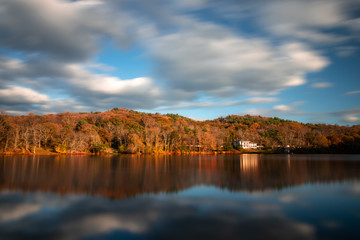 White puffy clouds streaking across the sky reflecting in a lake surrounded by fall foliage. 