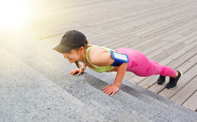 Cheerful fit woman in sportswear and cap doing push-ups from the stairs outdoors. Functional street workout.