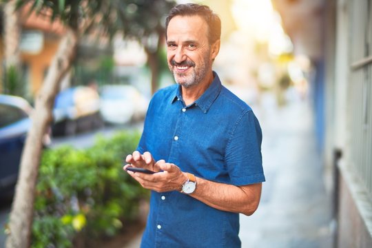 Middle age handsome businessman standing on the street using smartphone smiling