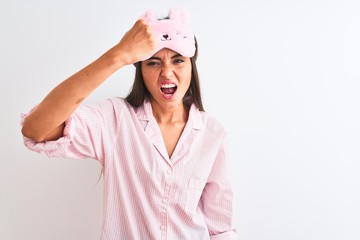 Young beautiful woman wearing sleep mask and pajama over isolated white background angry and mad raising fist frustrated and furious while shouting with anger. Rage and aggressive concept.