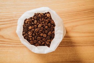 Roasted, brown coffee beans in white paper bag. Wooden background.