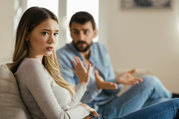 Worried young woman sitting on sofa at home and ignoring her boyfriend who is sitting next to her