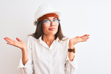 Young beautiful architect woman wearing helmet and glasses over isolated white background clueless and confused expression with arms and hands raised. Doubt concept.