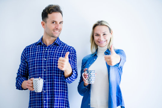 Young beautiful couple drinking cup of coffee standing over isolated white background happy with big smile doing ok sign, thumb up with fingers, excellent sign