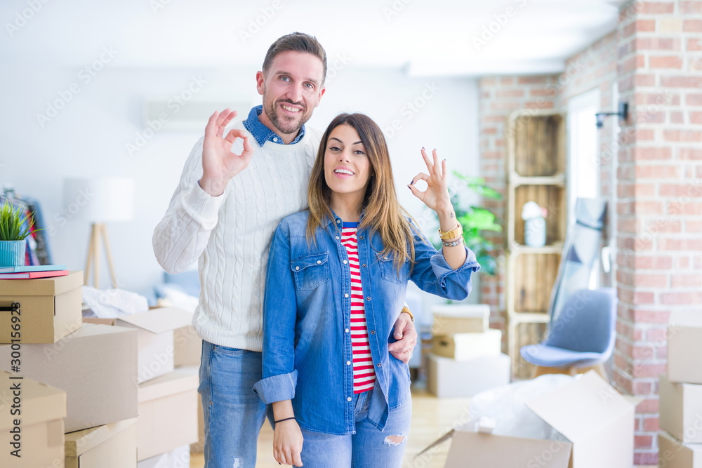 Sticker Young beautiful couple standing at new home around cardboard boxes smiling positive doing ok sign with hand and fingers. Successful expression.