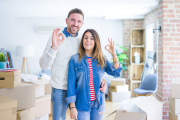 Obraz premium Young beautiful couple standing at new home around cardboard boxes smiling positive doing ok sign with hand and fingers. Successful expression.