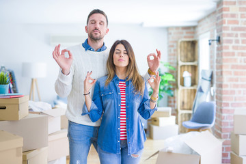 Young beautiful couple standing at new home around cardboard boxes relax and smiling with eyes closed doing meditation gesture with fingers. Yoga concept.