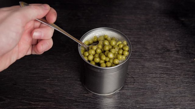 Male hand with spoon takes canned peas from tin can on black background.