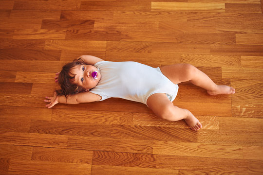 Beautiful toddler child girl wearing white bodysuit lying down on the floor using pacifier