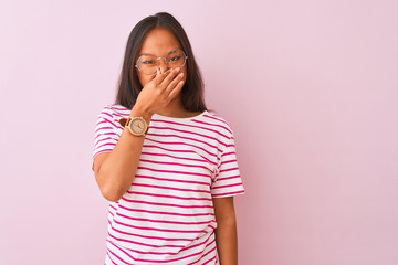 Young chinese woman wearing striped t-shirt and glasses over isolated pink background smelling something stinky and disgusting, intolerable smell, holding breath with fingers on nose. Bad smell