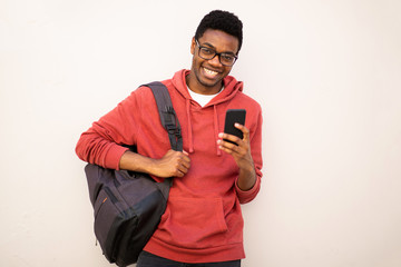 smiling young african american man with glasses and bag holding mobile phone by white background