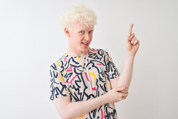 Young albino blond man wearing colorful t-shirt standing over isolated white background with a big smile on face, pointing with hand and finger to the side looking at the camera.