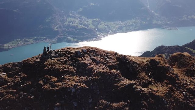 Drone shot of a hiker walking next to a summit cross on the stony top of a mountain next to a valley with a lake