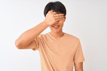 Young chinese man wearing casual striped t-shirt standing over isolated white background smiling and laughing with hand on face covering eyes for surprise. Blind concept.