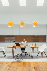 Concentrated busy caucasian male working online from home, sit thinking at table , white colors interior. Modern kitchen. Using laptop every day