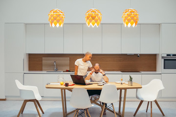 Concentrated man working at home using laptop. Sit at table in kitchen, woman support him stand next to man. White interior home