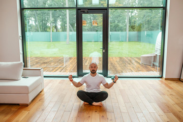 Male in casual clothes sit in lotus position meditate, relax in living room near sofa. Background...