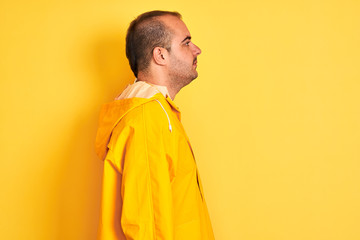Young man wearing rain coat standing over isolated yellow background looking to side, relax profile pose with natural face with confident smile.