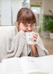 Young girl wrapping in plaid drinking thee at home, cold winter, reading book