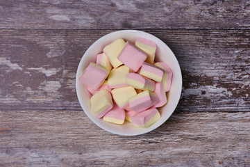 pink and yellow marshmallows in a white bowl in the middle of a wooden background, top view