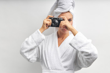 Caucasian pretty woman stand taking photo with photo camera, dressed in white bathrobe and towel on head. White isolated background