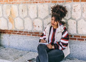 Fototapeta na wymiar Beautiful brunette girl with afro hair writes in her diary relaxingly while sitting next to a large stone and brick wall