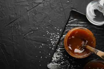 Glass jar with salted caramel and spoon on black background, copy space