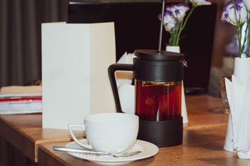 white cup on sauser, glass kettle with dark tea, empty paper blank with copy space and flowers on wooden table in cafe