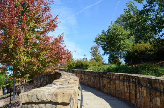 Looking up a winding walkway. Framed by a rock wall, the sidewalk is surround by fall colored trees.