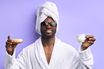 Handsome attractive man in towel and bathrobe holding cup of tea and donut, emotional. Cool boy in eyeglasses posing with donuts isolated over purple background